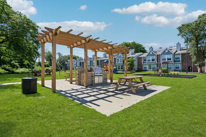 Charming outdoor area with BBQ grill, pergola, picnic tables, and lush green space, ideal for relaxation and gatherings.