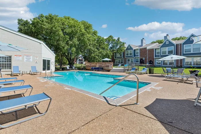 Inviting outdoor pool area with comfortable lounge chairs, surrounded by charming residential housing and lush greenery under a clear blue sky.