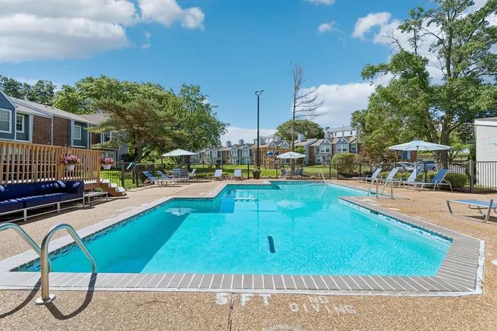 Inviting outdoor pool area surrounded by lush greenery and charming townhouses, featuring comfortable lounge chairs and umbrellas for relaxation.