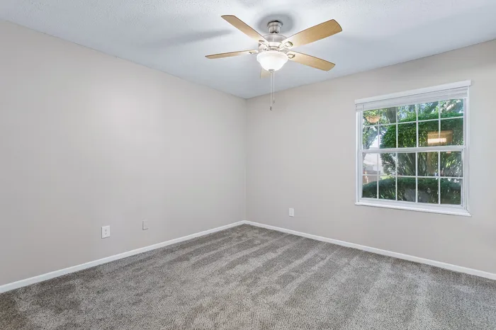 Bright and spacious bedroom featuring plush carpeting and a ceiling fan, with large window offering a view of lush greenery.