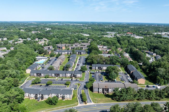 Aerial view of a serene apartment complex surrounded by lush greenery, featuring spacious layouts and ample parking, with a community pool and scenic suburban landscape.