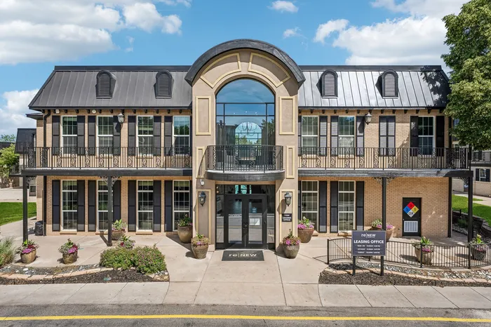 Elegant two-story brick building with large windows and a grand arched entrance, surrounded by landscaped gardens and vibrant flower pots.