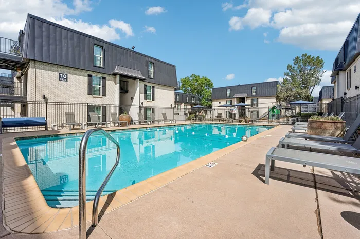 Inviting apartment pool area with modern lounge chairs, surrounded by charming brick buildings under a bright blue sky.