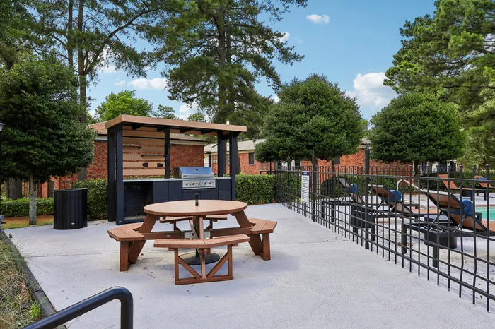 Charming outdoor picnic area with BBQ grill alongside a refreshing pool, surrounded by lush greenery and brick architecture.