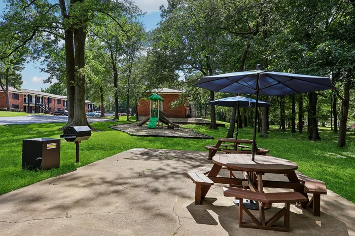 Inviting outdoor area with picnic tables and umbrellas, surrounded by lush greenery and a modern playground in the background.