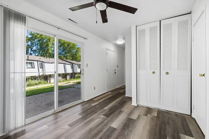 Bright and modern room with elegant wood flooring, large sliding glass door leading to a lush exterior view, and ample closet space under a ceiling fan.