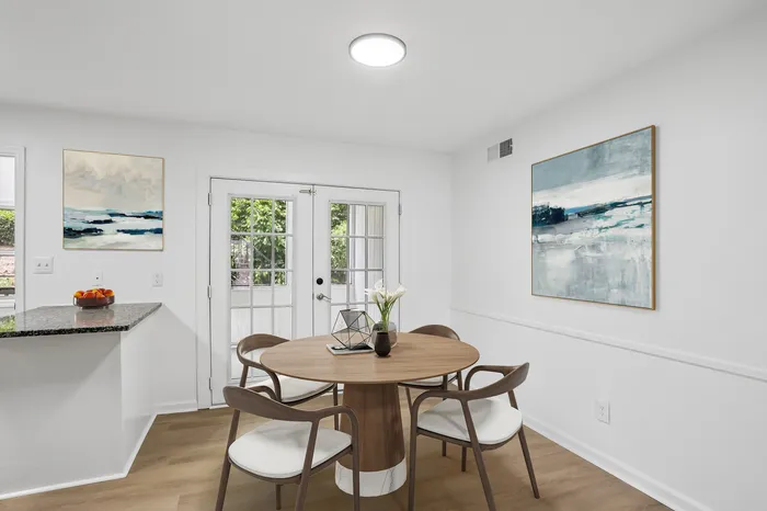 Bright dining area with elegant round wooden table, chic chairs, modern art, and natural light from French doors.