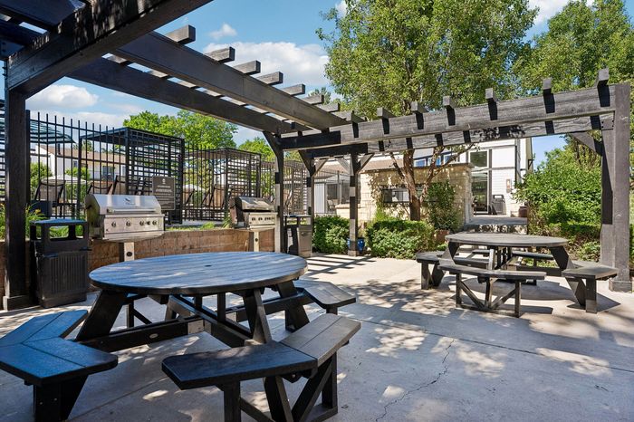 Charming outdoor patio with picnic tables and grills under a stylish pergola, surrounded by lush greenery for a perfect gathering spot.