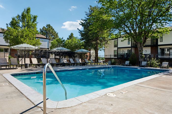 Inviting outdoor pool area with relaxing lounge chairs and sun umbrellas, surrounded by lush greenery and modern two-story buildings.