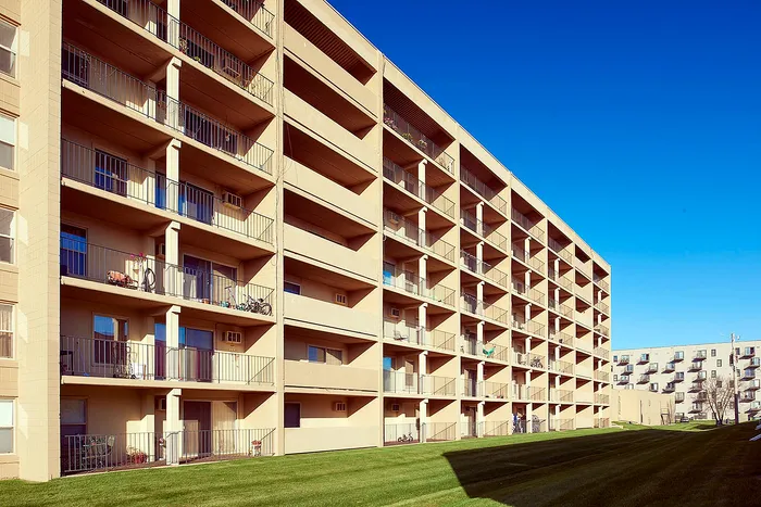 Sunlit apartment building with spacious balconies and lush green lawns, featuring modern architecture under a clear blue sky.