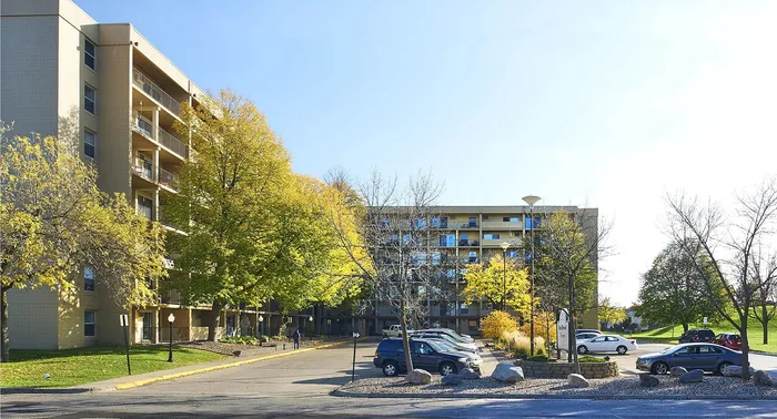 Sunny and inviting apartment complex surrounded by lush green trees and ample parking in a serene neighborhood setting.
