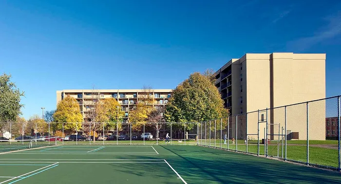 Bright apartment complex with spacious balconies and a lush green landscape, adjacent to tennis courts under a clear blue sky.