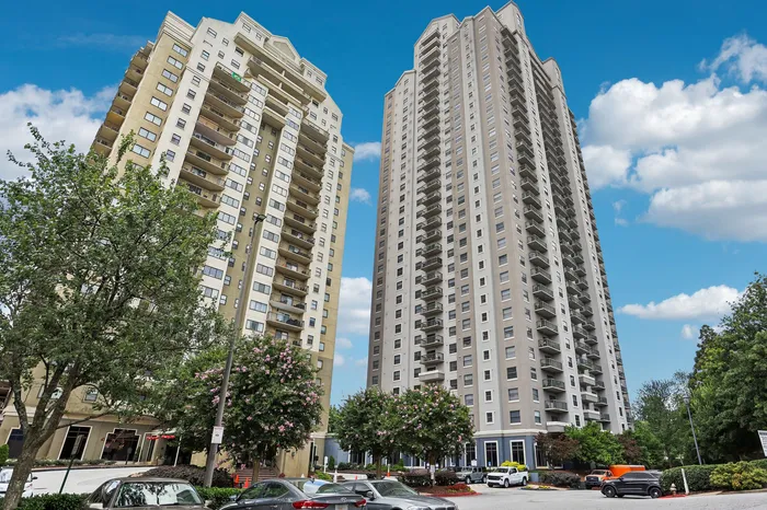 Modern high-rise apartment buildings with spacious balconies, surrounded by lush greenery and a vibrant urban setting under a clear sky.