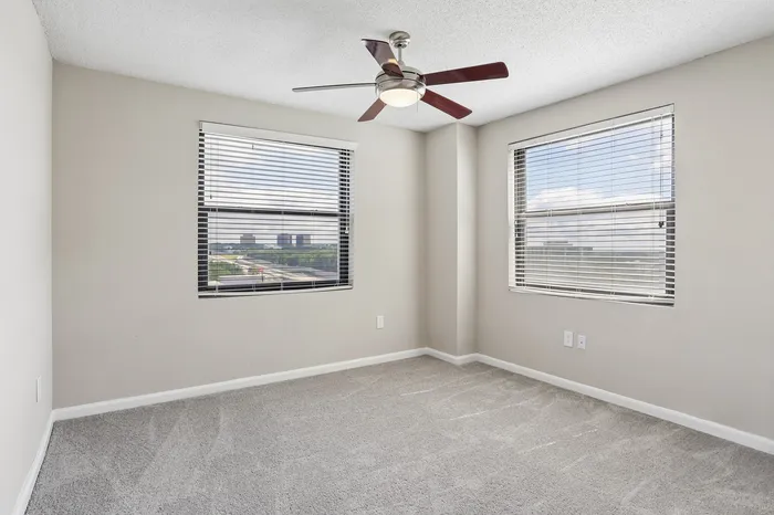 Bright and airy bedroom featuring plush carpet, neutral walls, and large windows with cityscape views, complete with a modern ceiling fan.