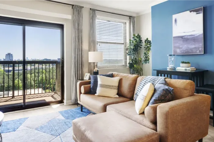 Bright living room with a stylish tan leather sofa, vibrant blue accents, and a stunning cityscape view through the balcony doors.