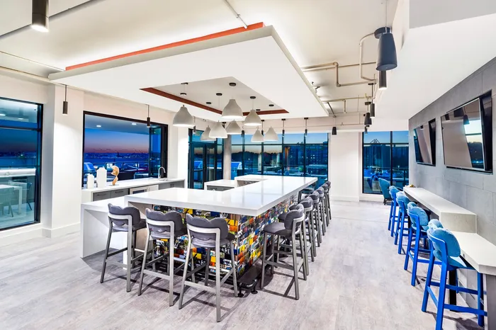 Modern communal kitchen with sleek countertops, vibrant barstools, and panoramic city views through large windows.