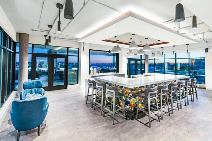 Modern communal lounge with a sleek island bar, blue armchair, and panoramic city views. Bright, spacious, and inviting atmosphere.