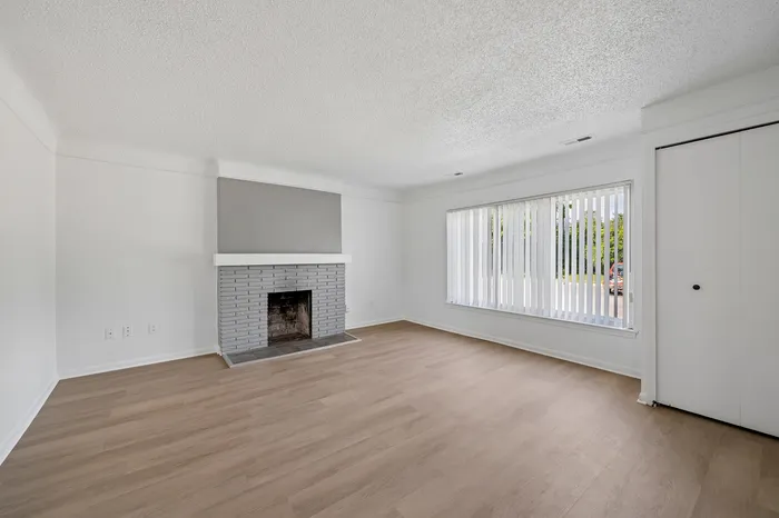 Bright, spacious living room with elegant hardwood floors, large window for natural light, and a stylish brick fireplace.