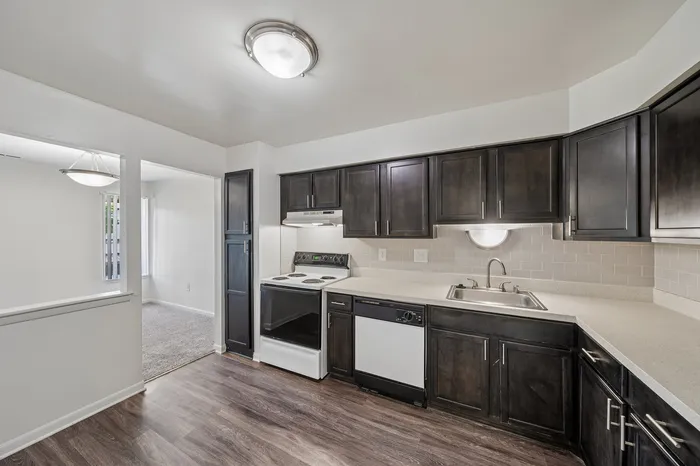 Modern kitchen with dark wood cabinets, sleek countertops, and stainless steel appliances, adjacent to a bright dining area with carpet flooring.