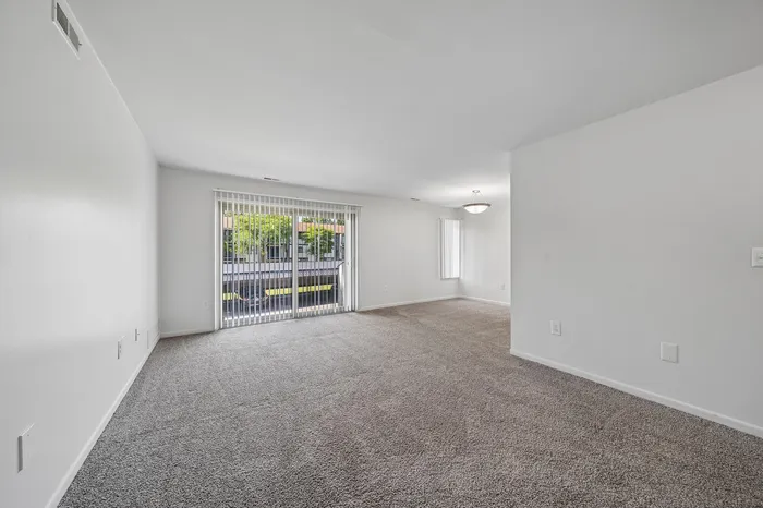 Bright and spacious open-plan living room with plush carpet and large sliding glass doors leading to a lush outdoor view.