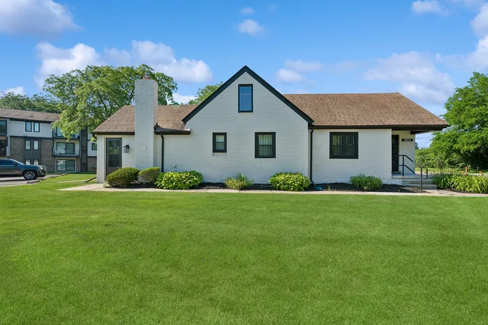 Charming white brick house with a gabled roof, surrounded by lush greenery and manicured lawns, under a bright blue sky.