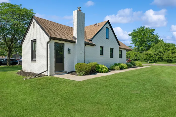 Charming white brick house with a gabled roof, lush green lawn, and vibrant plants, set against a backdrop of a clear blue sky.