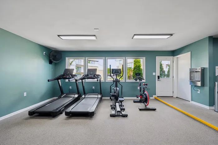 Modern fitness room with treadmills, exercise bike, and elliptical in a well-lit space featuring large windows and vibrant teal walls.