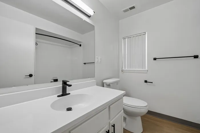 Modern bathroom with sleek black fixtures, spacious white vanity, and elegant wood flooring, complemented by natural light streaming through a window with blinds.