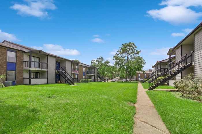 Charming apartment complex with private balconies, lush green courtyard, and inviting walkways under a clear blue sky.