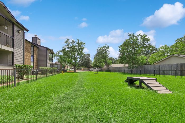 Lush green dog park with agility ramp, surrounded by charming apartment buildings under a bright blue sky.