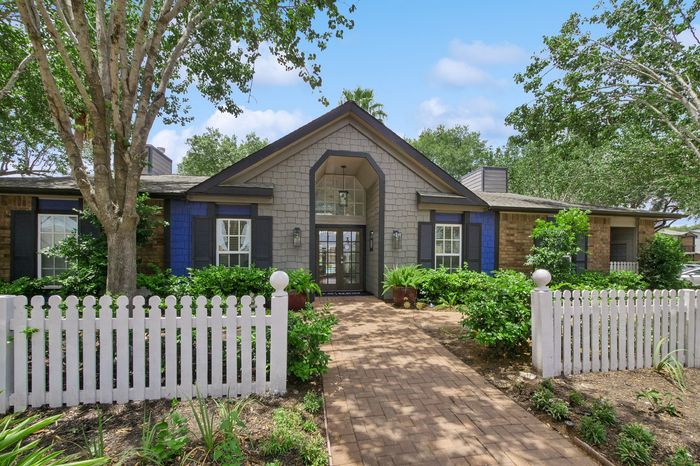 Charming residential home with a welcoming brick path, lush greenery, and a pristine white picket fence, set under a clear blue sky.