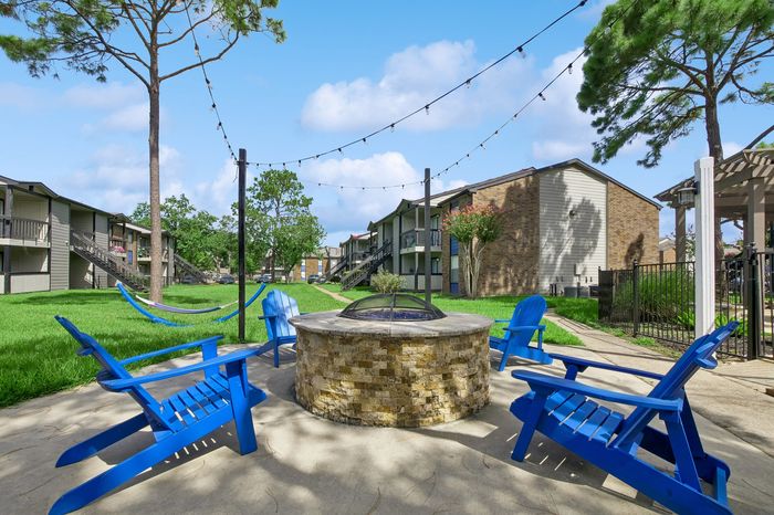 Vibrant outdoor space featuring a cozy fire pit surrounded by blue Adirondack chairs and hammocks, set amidst lush greenery under a blue sky.