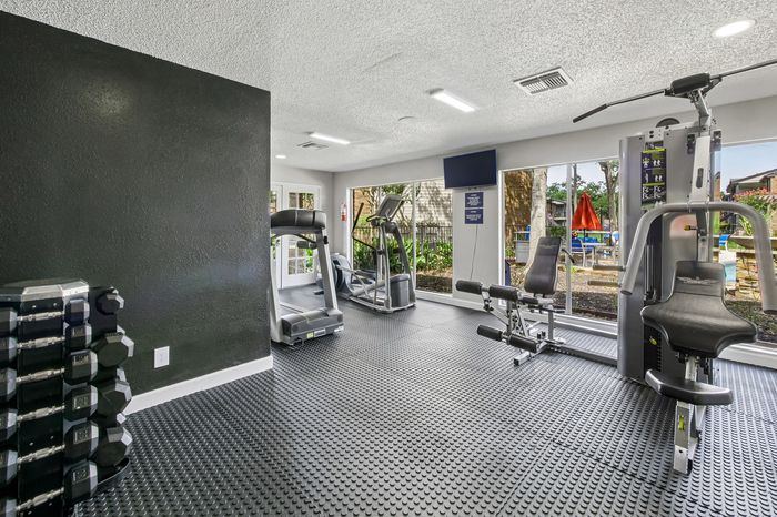 Modern apartment gym with state-of-the-art equipment, featuring treadmills, a weight station, and floor-to-ceiling windows offering a poolside view.