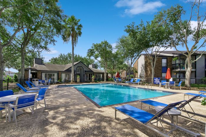 Inviting apartment complex boasting a crystal-clear swimming pool surrounded by lush greenery and modern lounging areas under a vibrant blue sky.