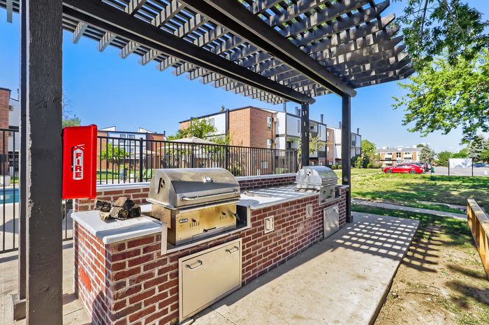 Modern outdoor barbecue area with sleek grills and brick structure, set in a vibrant apartment community with green lawns and contemporary buildings.