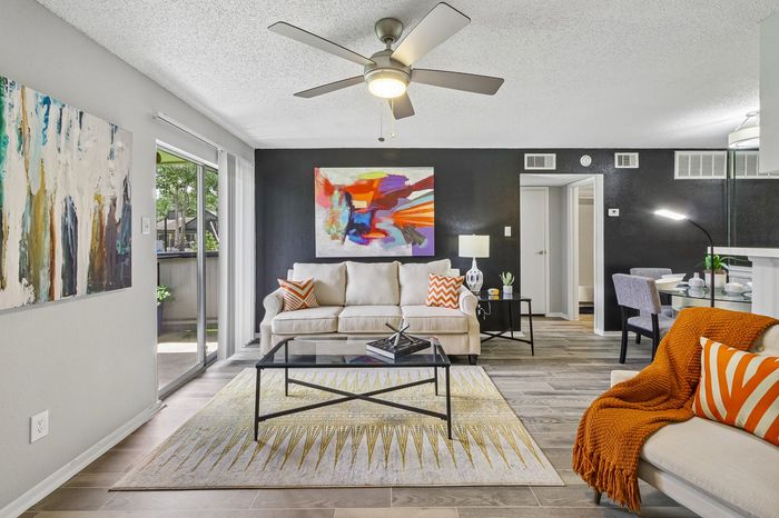 Modern living room featuring chic decor with abstract art, cozy beige sofas, and bright orange accents. Large window allows natural light over stylish flooring and a sleek glass coffee table.