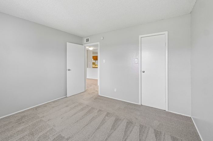 Bright, spacious bedroom with plush carpet and neutral paint, leading to a kitchen with wooden cabinetry.