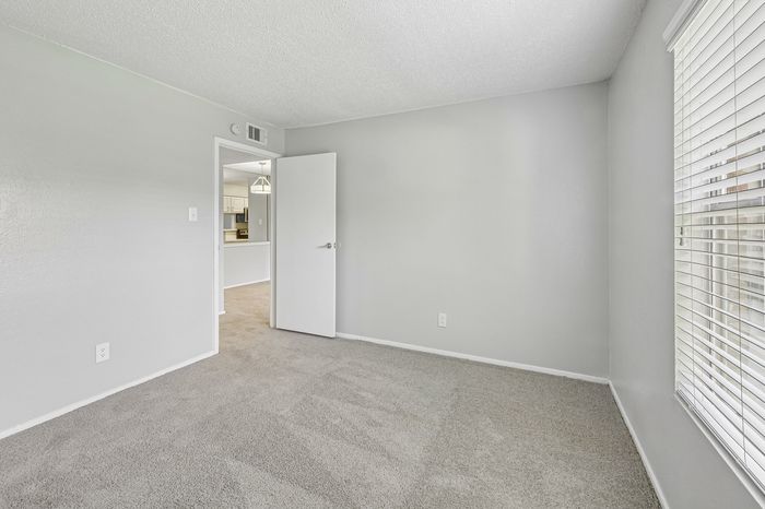 Bright and welcoming room with plush carpet, inviting natural light from large windows, and a view into the modern kitchen, perfect for comfortable living.
