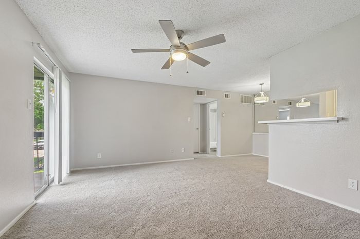Bright, open-concept living area with plush carpet, featuring a modern ceiling fan and sliding glass doors leading to a private patio.