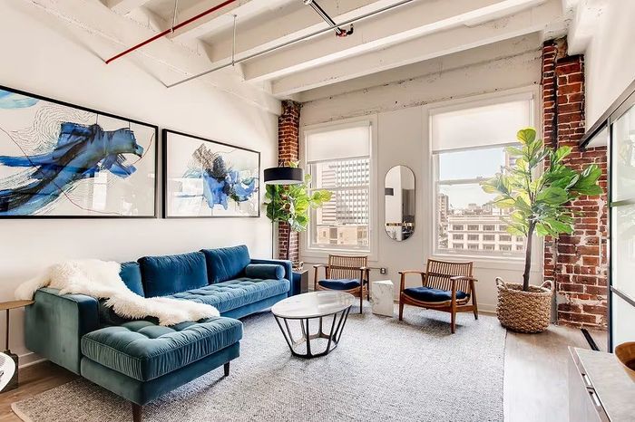 Chic urban living room with exposed brick, large windows, stylish blue sofa, modern art, and lush greenery.