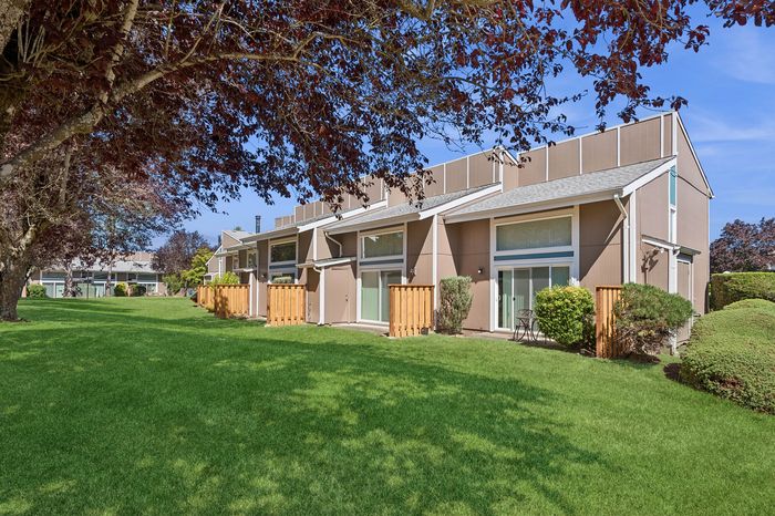 Charming townhouse units with large windows and private patios set against a lush, green landscape with mature trees.