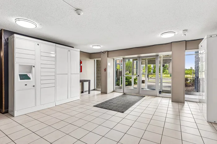 Bright and modern apartment lobby with stylish tile flooring, secure white parcel lockers, and large glass doors leading to a lush outdoor garden area.