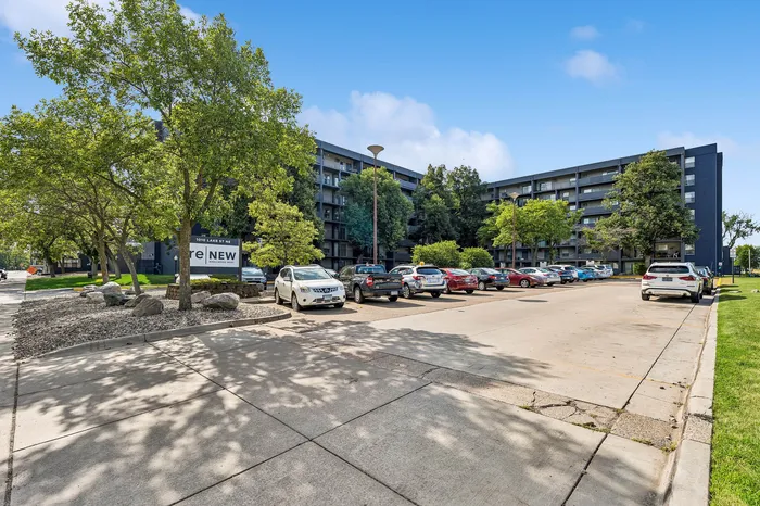 Modern apartment complex with abundant parking, surrounded by lush greenery and clear blue skies.