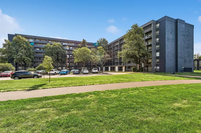 Modern apartment complex with lush green surroundings and ample parking under a clear blue sky, offering urban living in a serene environment.