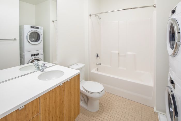 Modern bathroom with sleek white fixtures, a compact stacked washer and dryer, and natural wood cabinetry, offering convenience and style.