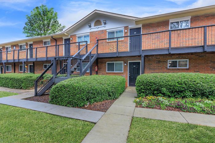 Charming brick apartment complex with modern balconies, lush greenery, and inviting walkways set under a clear blue sky.