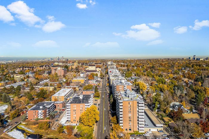 Aerial view of a vibrant urban neighborhood with tree-lined streets, modern apartment buildings, and a scenic city skyline in the background.