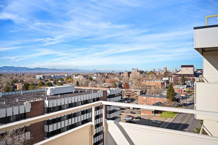 Scenic cityscape view from a modern apartment balcony, showcasing urban architecture against a backdrop of distant mountains and a clear blue sky.