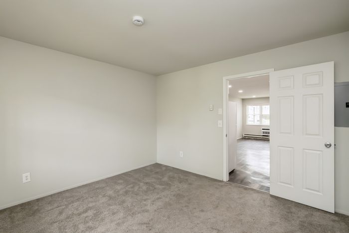 Inviting apartment interior showcasing a cozy bedroom with plush carpet and an open doorway leading to a well-lit living area with large windows.