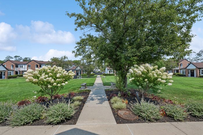 Charming apartment courtyard with lush green lawns, blooming flowers, and a serene walkway lined with trees and benches.